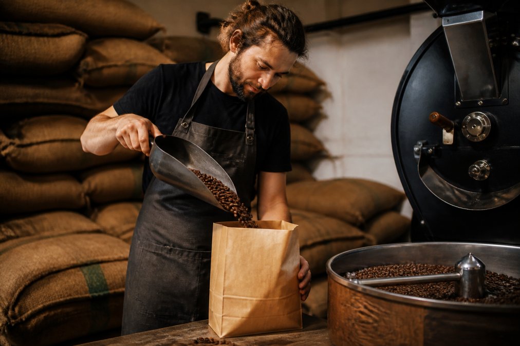 A roaster pouring freshly roasted whole bean coffee into a craft paper bag at a small roastery, warm lighting, burlap sacks stacked in the background