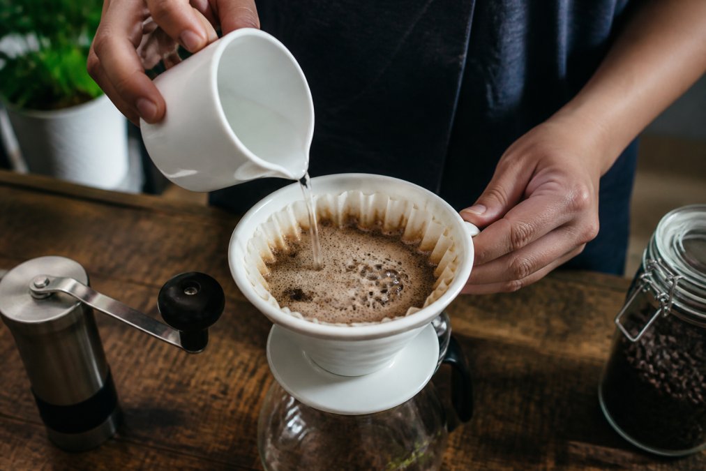 Close-up of a person's hands holding a small white ceramic cup over a pour-over dripper, watching coffee bloom and bubble as hot water hits freshly ground coffee beans on a wooden kitchen counter