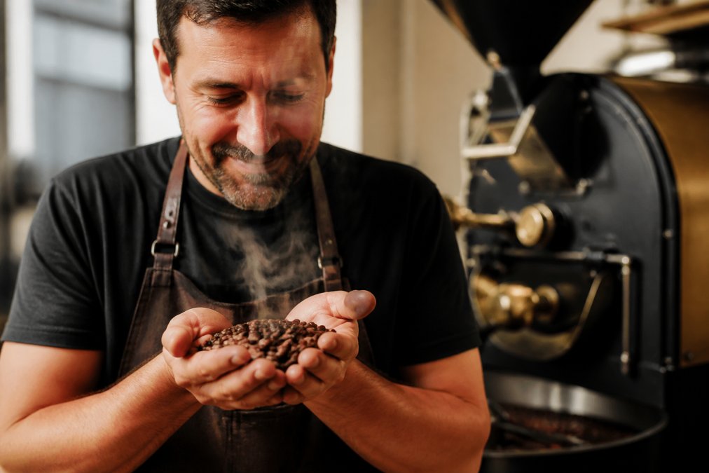 Close-up of freshly roasted coffee beans cupped in a person's hand, gentle steam rising, with a small-batch roaster drum softly blurred in the background of a warmly lit roastery