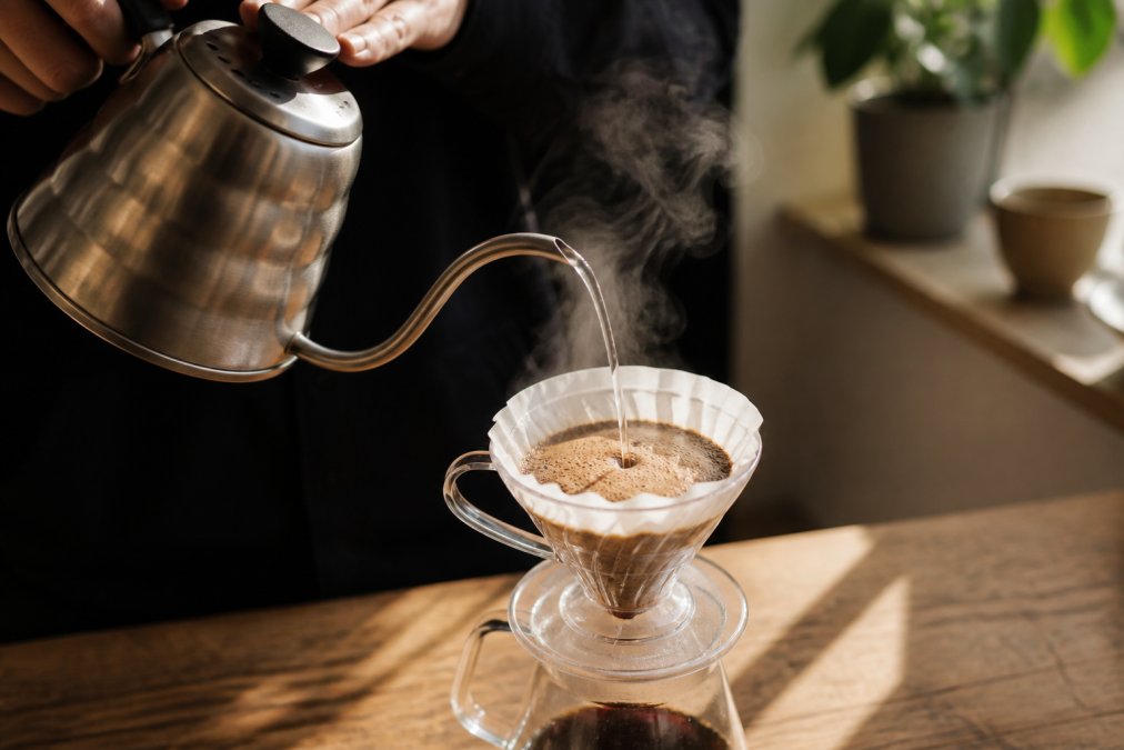Close-up of hands pouring hot water over coffee grounds in a pour-over cone setup, steam rising visibly, showing the blooming phase of brewing with natural morning light